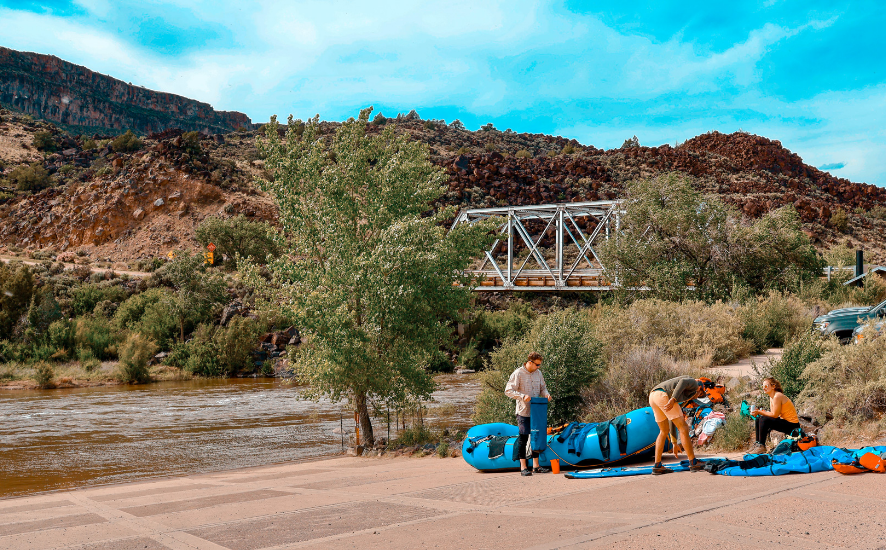 Rafters prepare boats at river's edge near bridge